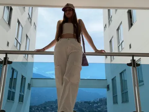 Young woman in white linen trousers and crop top standing on a modern balcony overlooking a city view.