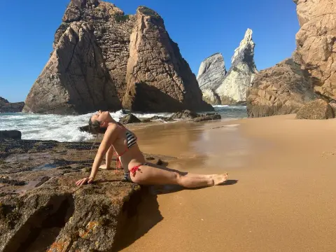 Huanshulann wearing a striped bikini, posing on a rock at the beach with ocean waves and large cliffs in the background.