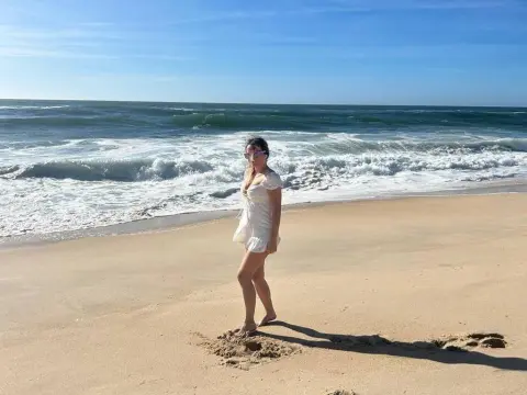 Huanshulann stands on a sandy beach wearing a short white dress, looking back over her shoulder with the ocean waves behind her.