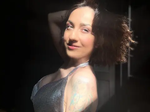 Smiling woman in a silver sequin top enjoying warm sunlight in a moody, dark indoor setting.