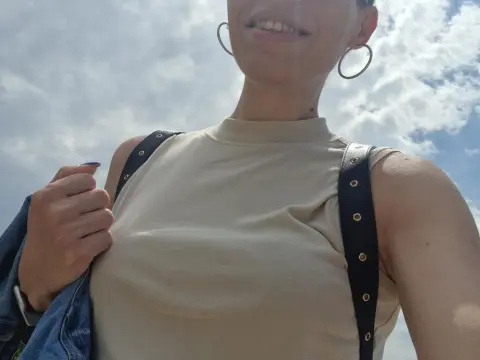 QueenOfLust — Smiling woman in a beige sleeveless top and hoop earrings enjoying a sunny day under a blue sky.