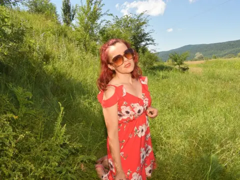 KateConner poses in a red floral sundress and sunglasses while standing in a grassy field with green trees and hills in the background.