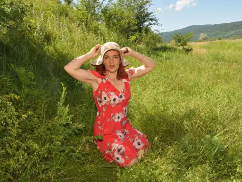 KateConner poses in a red floral sundress and a straw hat while kneeling in a grassy field under a clear blue sky.