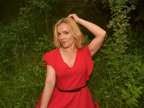 KateConner poses in a red dress with her hand in her blonde hair while standing in a grassy, wooded area.