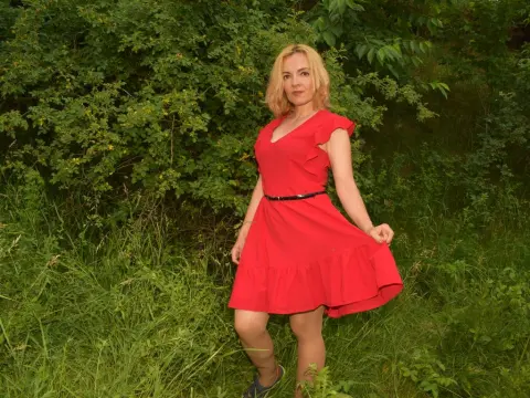 KateConner wearing a short red dress while standing in a grassy, leafy outdoor setting.