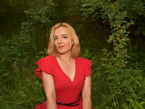 KateConner poses in a red dress with a v-neckline, standing against a background of thick green leaves and bushes.