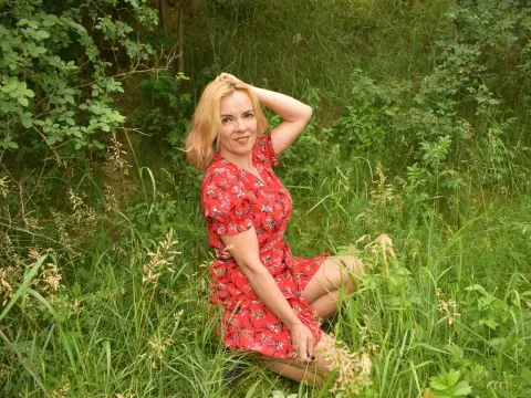 KateConner poses in a red floral dress while kneeling in tall green grass, running her hand through her blonde hair.