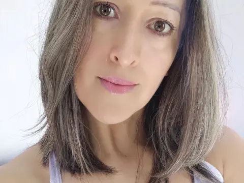 Gaby_Sexy — Close-up portrait of a smiling woman with long grey hair wearing a white tank top in a bright indoor setting.