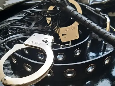 NikkiMistress — Silver metal handcuffs and a black studded leather belt on a dark textured surface for a bold fetish aesthetic.