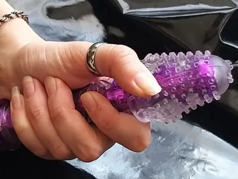 NikkiMistress — A person's hand holding a textured purple silicone massager against a black satin fabric background.