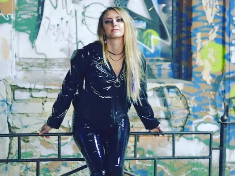 NikkiMistress — Edgy blonde woman in black patent leather outfit posing against a colorful graffiti wall in an urban setting.