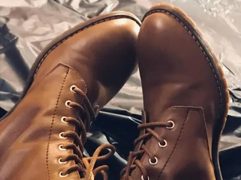 NikkiMistress — Close-up of stylish brown leather lace-up combat boots resting on a textured fabric background.