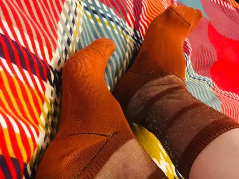 NikkiMistress — Close-up of feet wearing cozy burnt orange patterned socks resting on a colorful striped blanket.