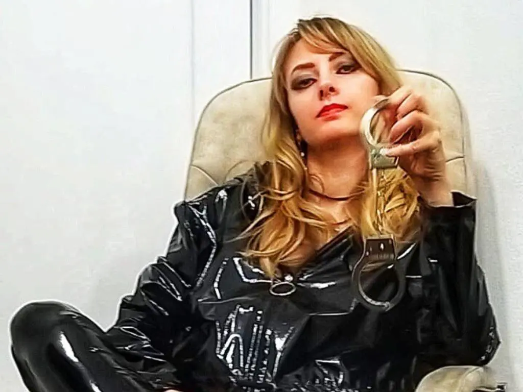 NikkiMistress — Blonde woman in black latex outfit holding handcuffs while sitting in a chair with a moody expression.