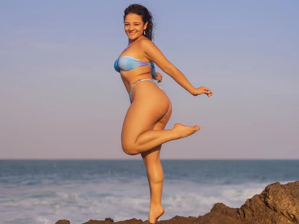Lilly_boss — Smiling woman in a blue bikini posing joyfully on rocky coastal cliffs by the ocean at sunset.