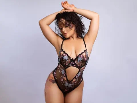Curvy woman in black floral lace bodysuit posing confidently against a plain studio background.