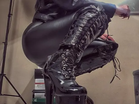 Woman in black leather outfit and high lace-up patent platform boots sitting in a studio setting.