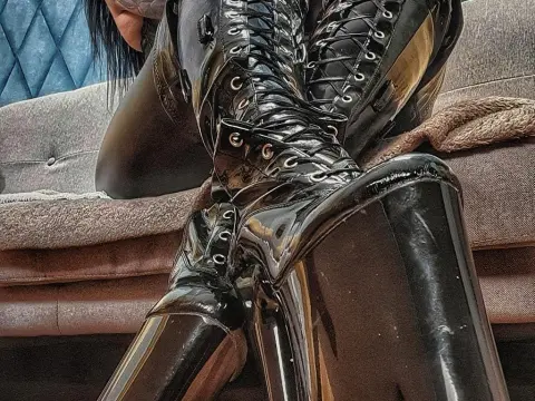 Woman in shiny black latex bodysuit and lace-up thigh-high boots sitting on a sofa in a moody setting.