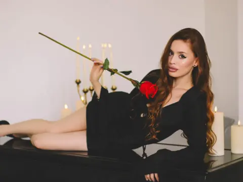 CherylJess poses reclining on a black surface, wearing a black long-sleeve top and holding a single long-stemmed red rose near her face.