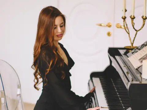 CherylJess with long wavy brown hair, wearing a black blazer while sitting at a black piano in a bright room.