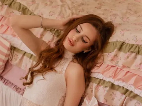 CherylJess lies on a floral bedspread with her long brown hair spread out and one hand resting behind her head, wearing a white lace top.