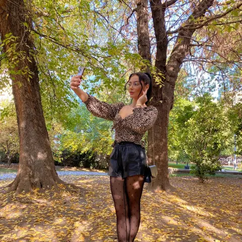 AngieShae — Woman in leopard print blouse and black shorts taking a selfie in a sunny autumn park with fallen leaves.