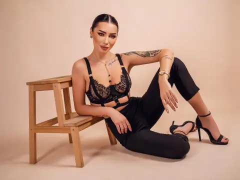 AngieShae — Tattooed woman in black lace lingerie and trousers sitting on a wooden stool against a beige studio background.