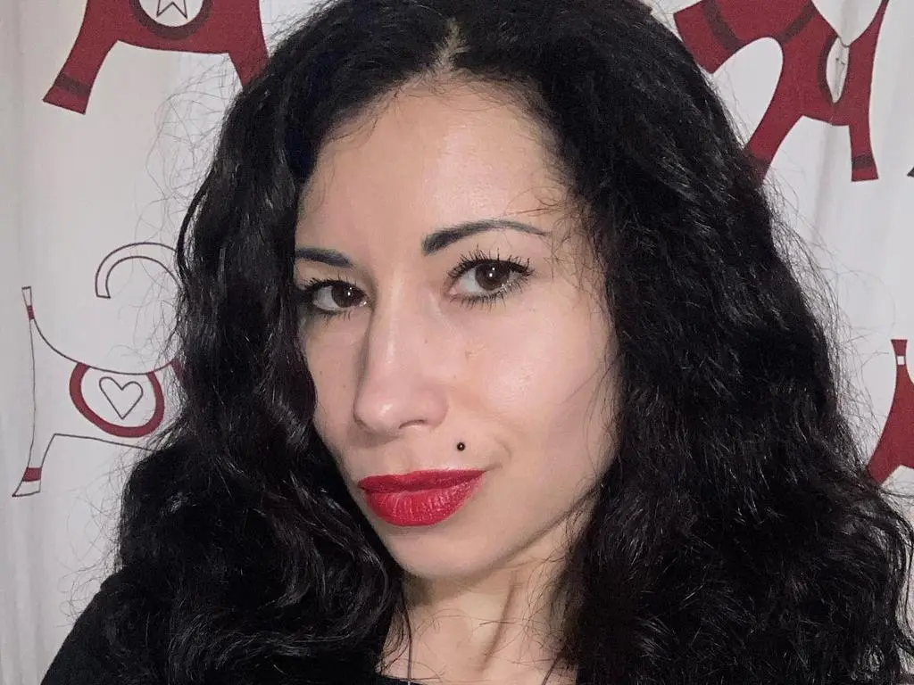 XLittleMistressX — XLittleMistressX poses with her long, wavy black hair and bright red lipstick against a white patterned background.