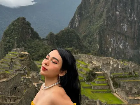 Mindy_Star — Mindy_Star poses with eyes closed, wearing a white pearl necklace and a yellow top against the stone ruins and green mountains of Machu Picchu.