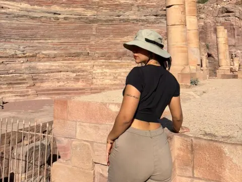 Mindy_Star — Mindy_Star poses outdoors in a black t-shirt and tan trousers, wearing a wide-brimmed sun hat while looking away from the camera.