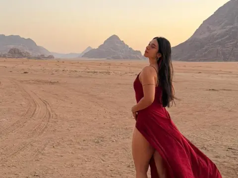 Mindy_Star — Mindy_Star poses in a long red dress with a high slit, standing in a desert landscape with mountains behind her.