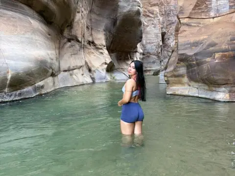 Mindy_Star — Mindy_Star stands waist-deep in a narrow canyon river, wearing a white bikini top and blue shorts with her long dark hair down.