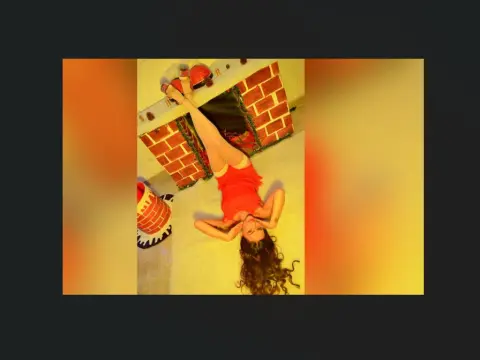 Young woman in a red summer dress relaxing on a floor near a brick fireplace in a warm, sunlit room.