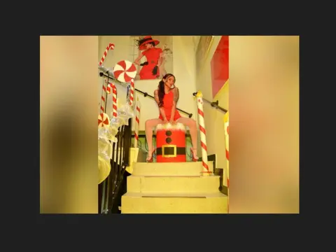 Woman in a red festive outfit sitting on a giant candy prop in a bright, whimsical holiday setting.