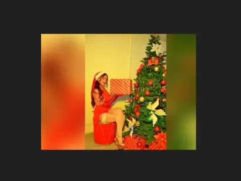 Smiling woman in a red Santa outfit holding a gift next to a decorated Christmas tree in a festive holiday setting.