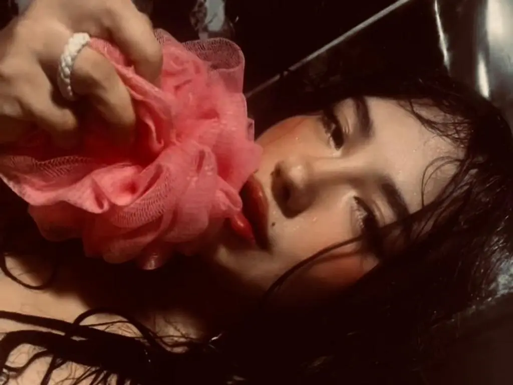 Perla_Dumon holds a pink mesh bath sponge against her lips while lying in water with wet, dark hair.