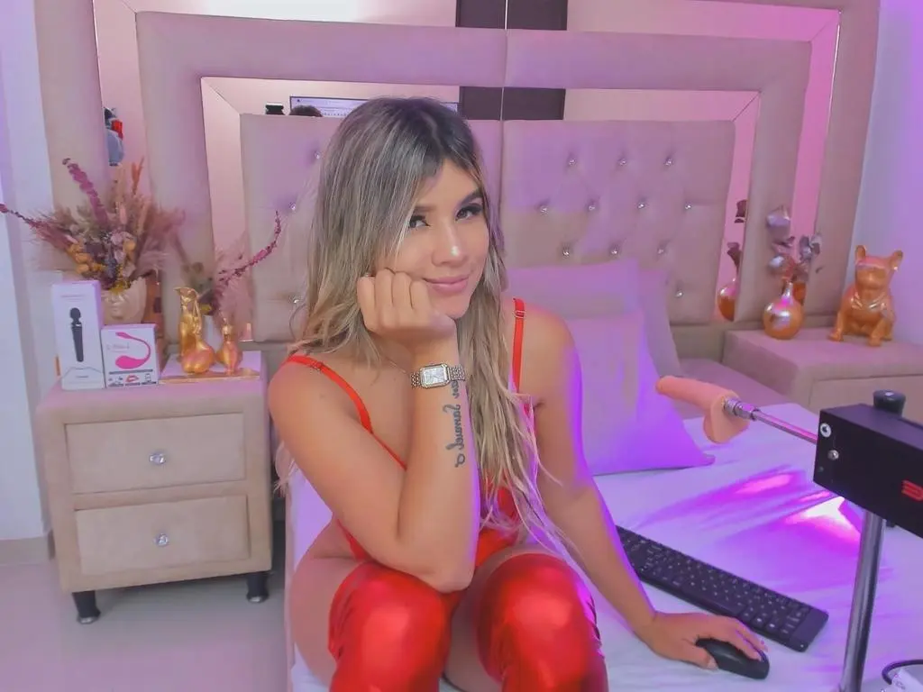 Karla_Ross — Smiling woman in a red outfit sitting on a bed in a bright, modern bedroom setting.