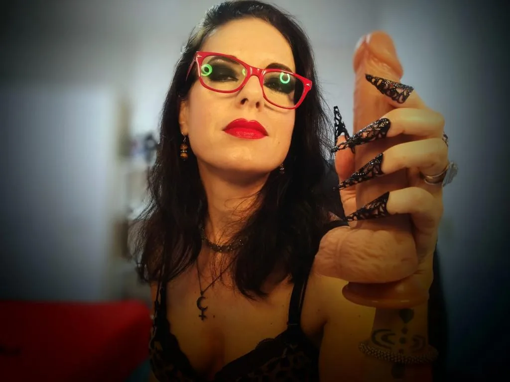 MrsBlackMoonLilith wears red glasses and black lace while holding a large flesh-colored dildo toward the camera.