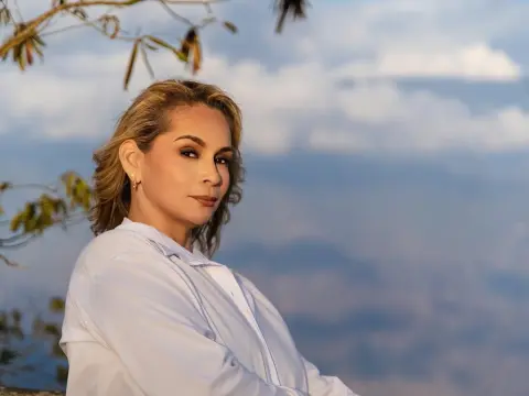 Smiling blonde woman in a white button-down shirt posing against a soft blue sky at sunset.