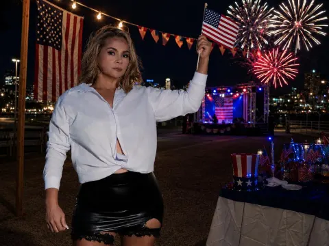 CarlaOlivieri — CarlaOlivieri wears a white button-down shirt and black leather mini skirt while holding a small American flag at night with fireworks in the background.