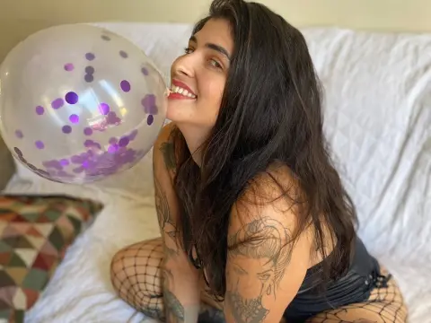 Smiling woman with tattoos blowing a clear polka dot balloon while sitting on a white bed in a cozy room.