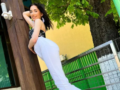 Smiling woman in a black patterned top and white trousers leaning against a wooden pillar in a sunny garden.