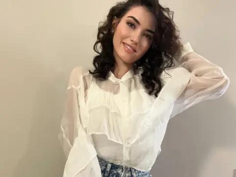 Smiling woman with curly hair wearing a white sheer ruffled blouse in a minimalist indoor setting.