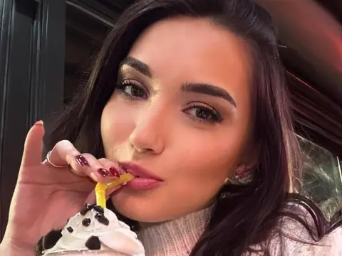 Smiling woman with dark hair sipping a milkshake through a straw in a cozy indoor setting.