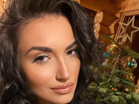 Beautiful woman with wavy dark hair smiling in front of a decorated Christmas tree in a cozy wooden cabin.