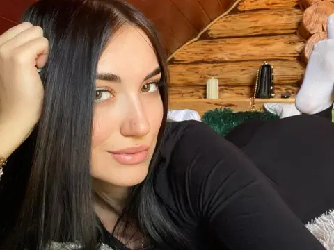 Smiling woman with long dark hair wearing a black long-sleeve top in a cozy wooden cabin setting.