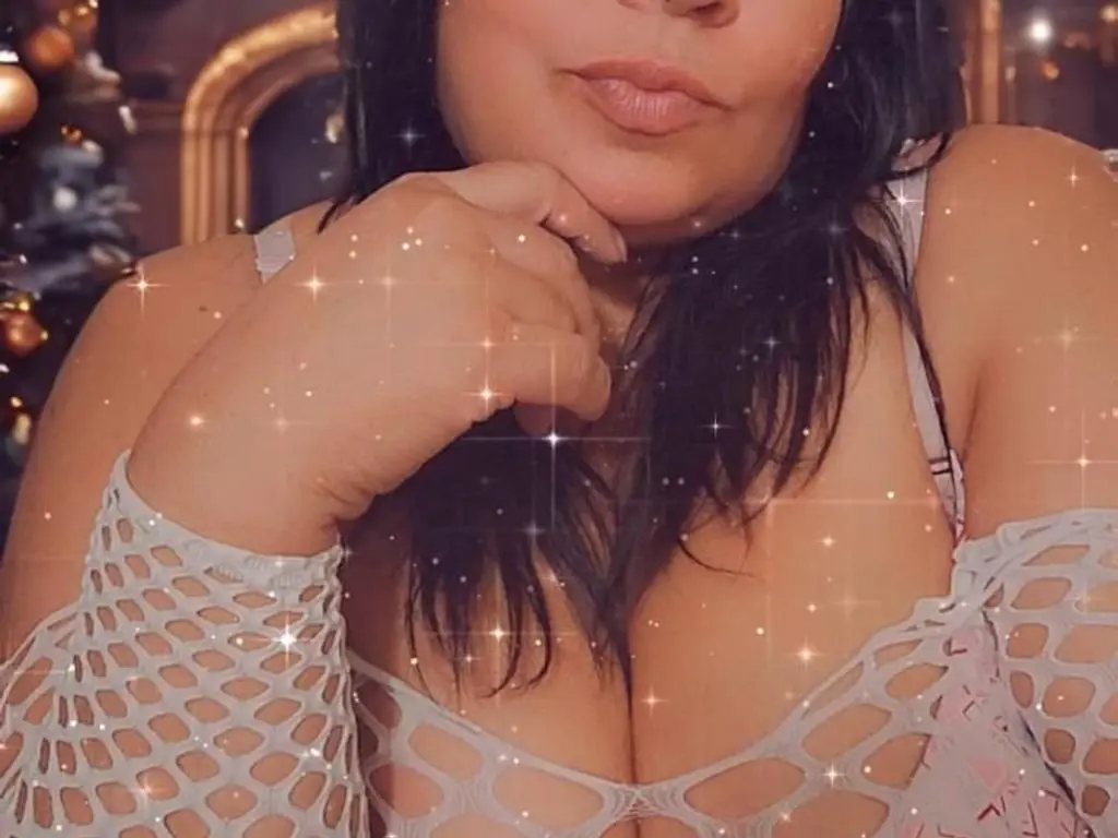NICOLDAYANA69 poses with her hand under her chin, wearing a white fishnet top that shows her cleavage and shoulders.