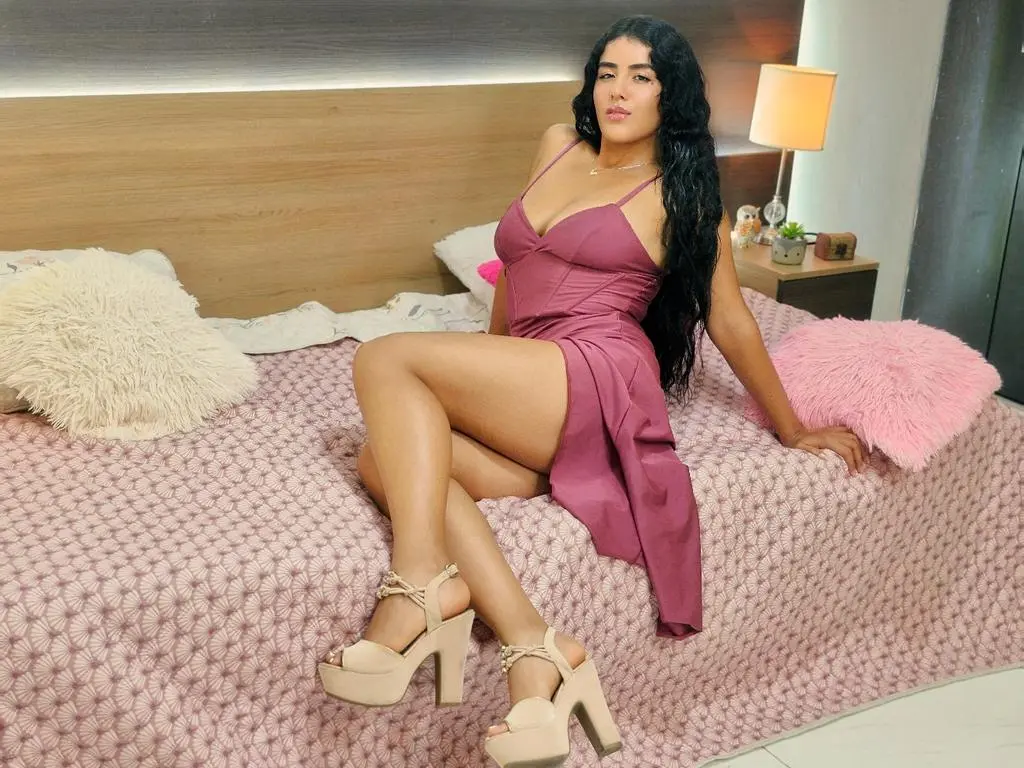Flower_Cute — Woman in a mauve satin dress and beige platform heels sitting on a pink bed in a cozy bedroom setting.