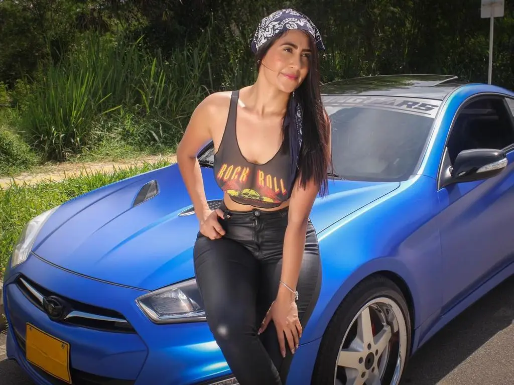 AntonelaDreamss — Stylish woman in black tank top and jeans posing confidently next to a blue matte sports car outdoors.