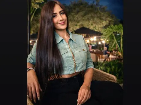 Smiling woman in a light denim jacket and black pants sitting outdoors at a tropical evening lounge.